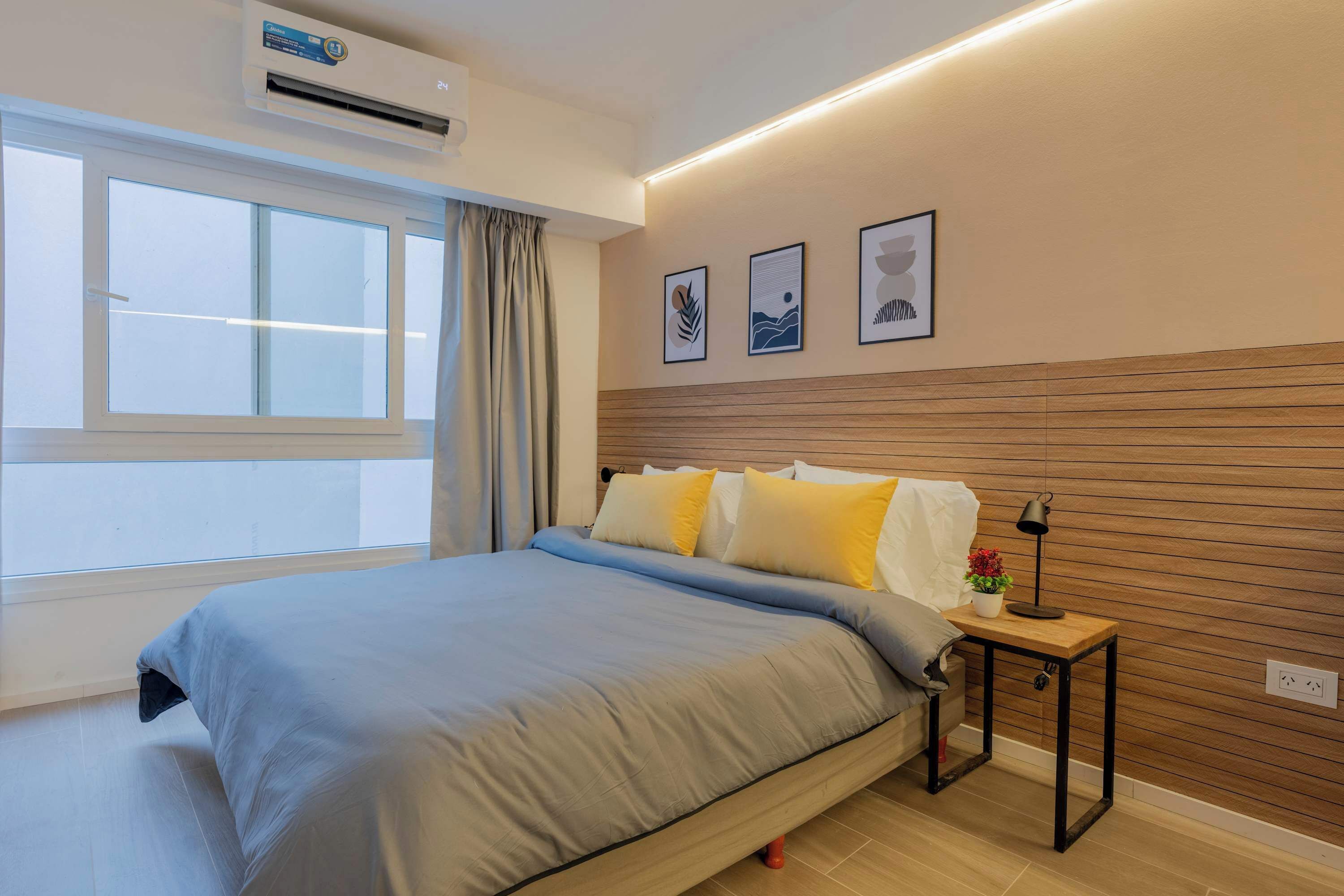 Un dormitorio con cama, mesita de noche con lámpara, ventana y una obra de arte en la pared.