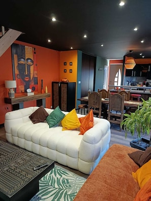 A modern living room with a white sofa, colorful cushions, a patterned rug, and a dining area with wooden chairs and a table.