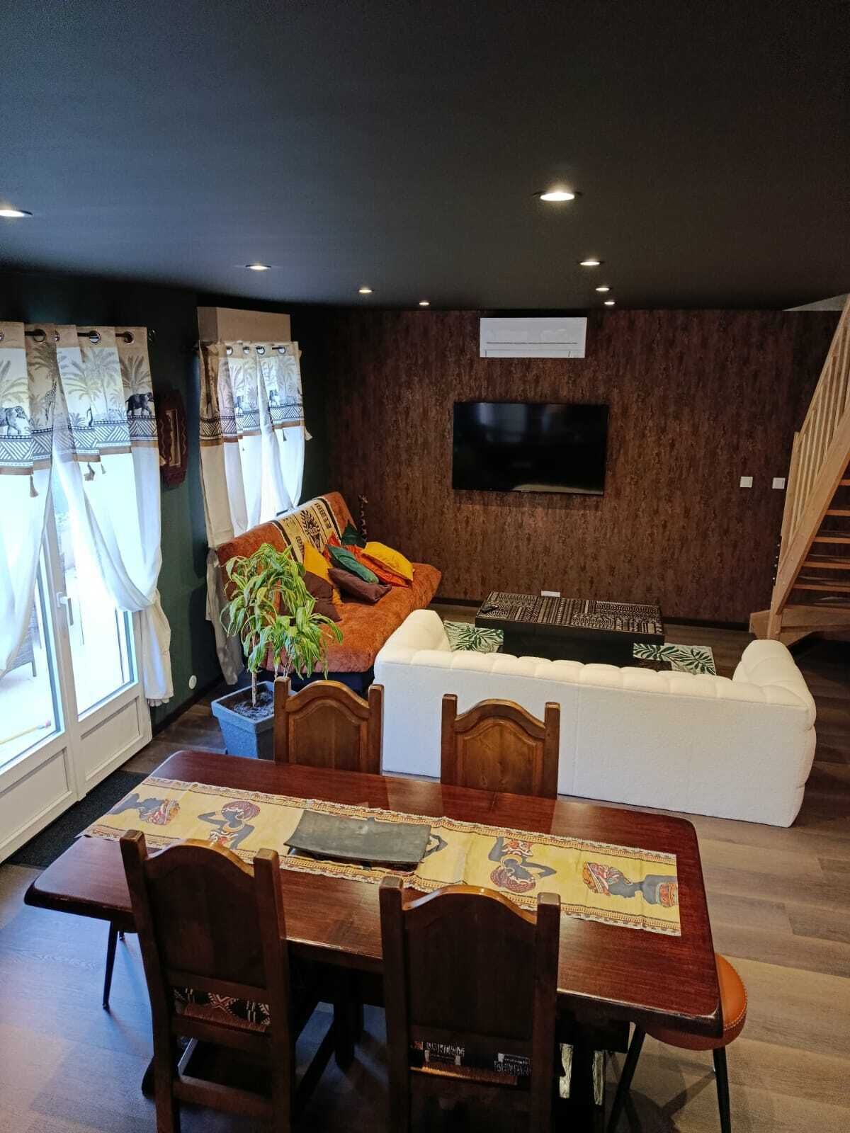 Un coin repas avec une table et des chaises en bois, un canapé, un téléviseur fixé au mur et un escalier en arrière-plan.