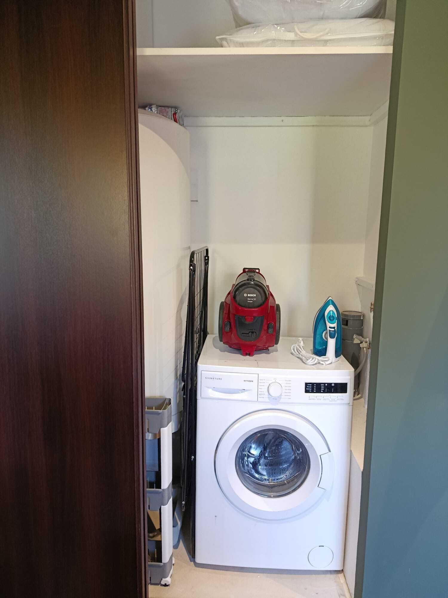 A laundry area with a washing machine, a red Bosch vacuum cleaner, and a blue iron on top.