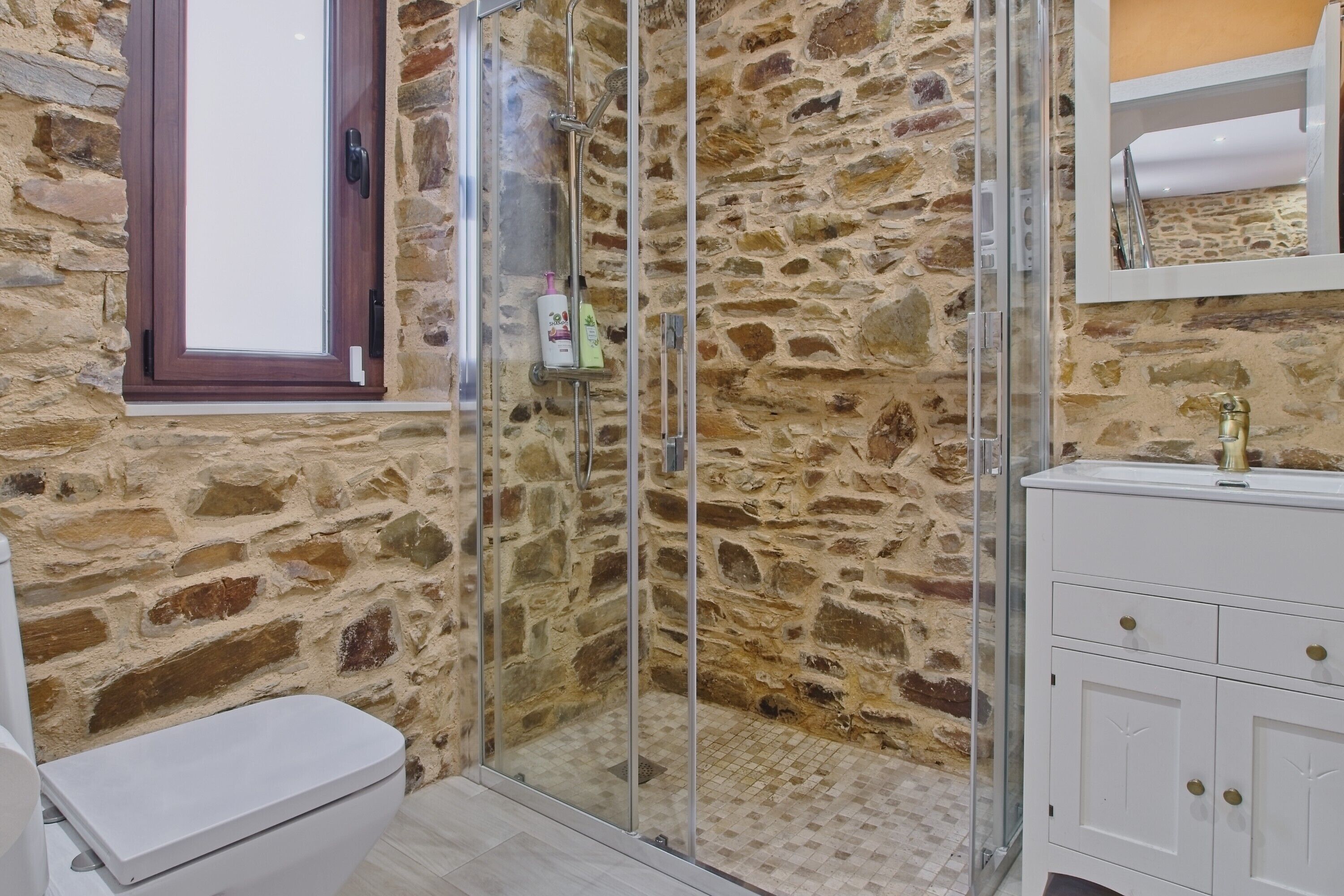 Un baño con pared de piedra, cabina de ducha de vidrio, inodoro blanco y tocador blanco con tiradores dorados.