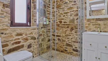 Un baño con pared de piedra, cabina de ducha de vidrio, inodoro blanco y tocador blanco con tiradores dorados.
