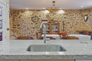 A stone-walled room with a granite countertop sink, wooden dining table, chairs, and a chandelier.