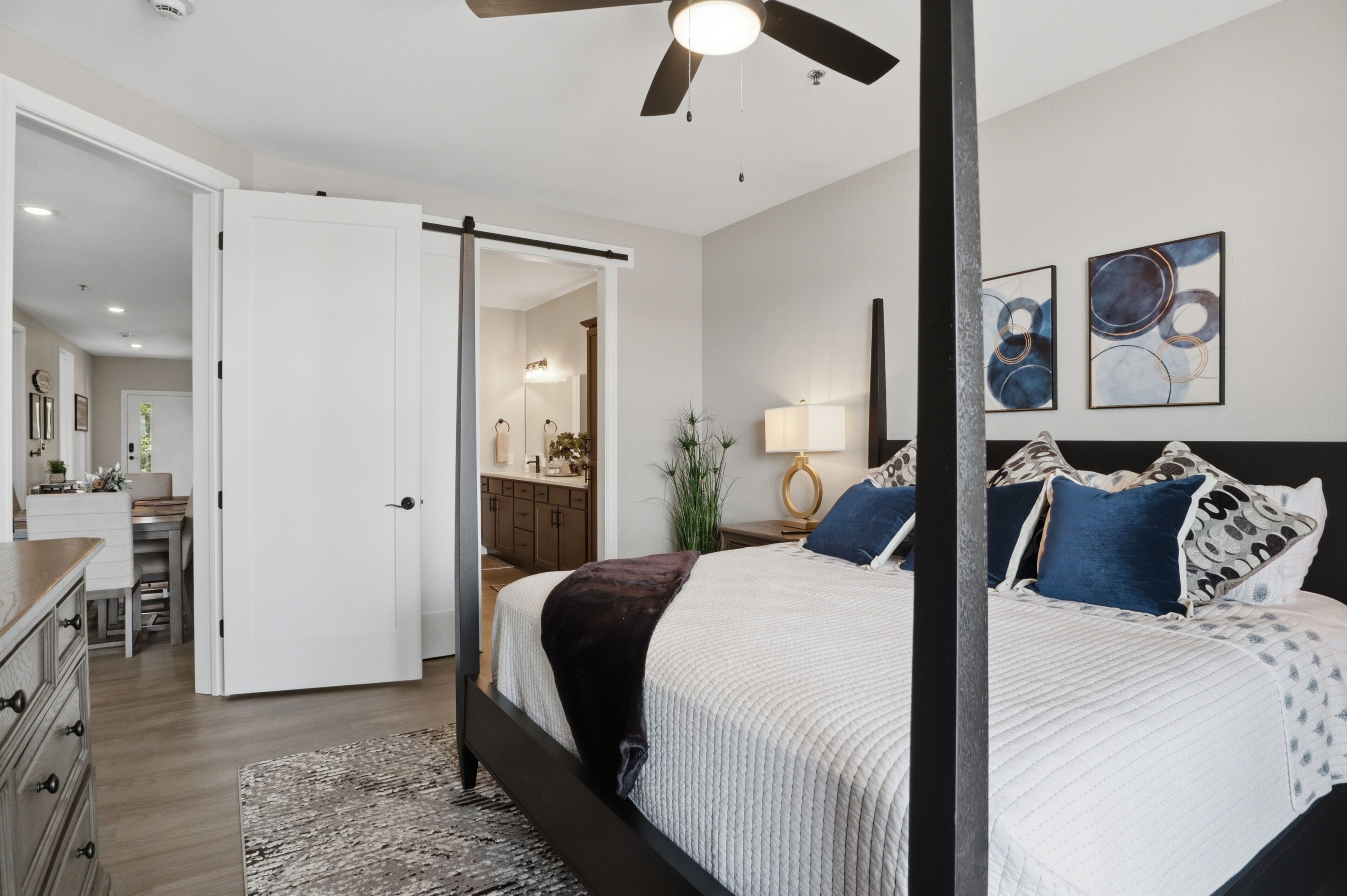 A bedroom with a bed, a ceiling fan, a nightstand, a lamp, and artwork on the wall.