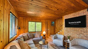 Una habitación acogedora con paredes de madera, un muro de acento de ladrillo, un sofá, dos sillones, una mesita con una lámpara y un televisor montado en el muro de ladrillo.