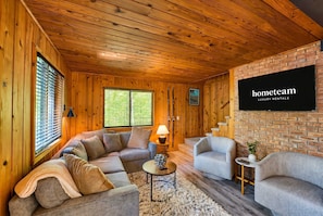 A cozy room with wooden walls, a brick accent wall, a sofa, two armchairs, a small table with a lamp, and a TV mounted on the brick wall.