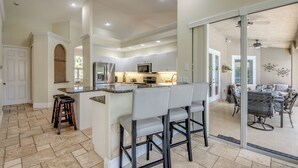 A modern kitchen with a central island, bar stools, and a dining area with a glass door leading to a patio. - Heated Pool - Spacious - Bike - BBQ- Sound System (Cape Coral)