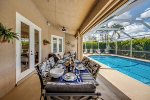 Un área de comedor junto a la piscina, con una mesa dispuesta para cuatro personas, sillas alrededor y vista a una piscina.