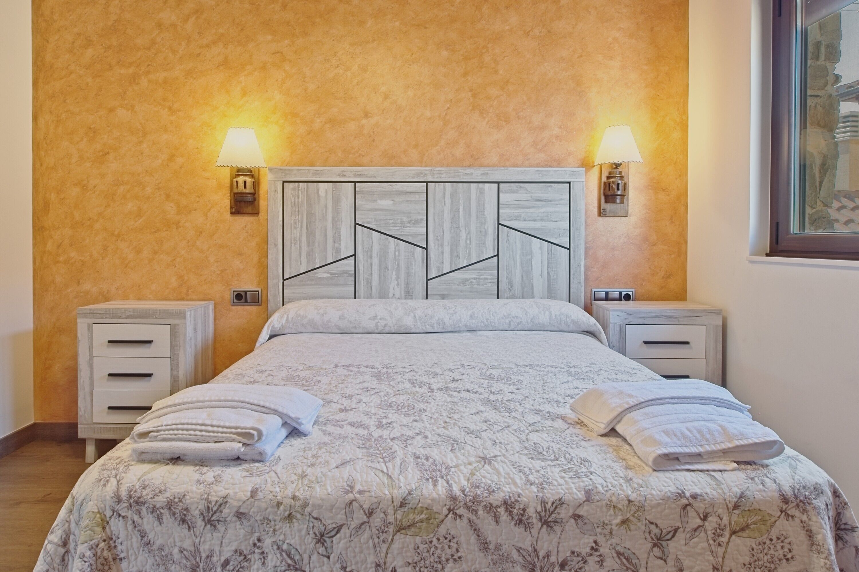 A bedroom with a wooden headboard, two bedside tables with lamps, a floral bedspread, and a neatly made bed with towels.