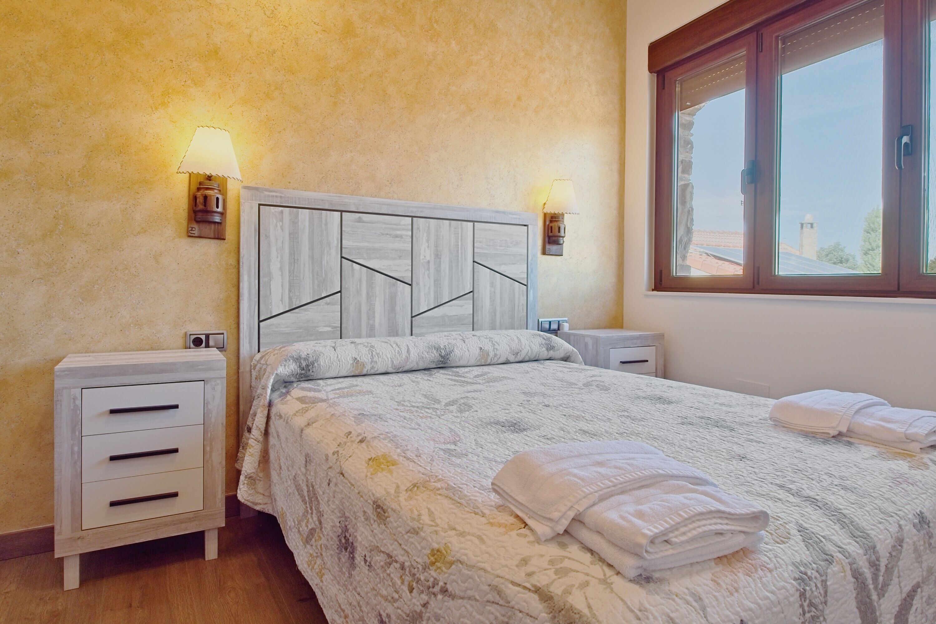 Un dormitorio con cabecera de madera, dos mesitas de noche, una cama con colcha estampada, dos lámparas de pared y un ventanal con vistas.