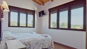 Quarto com cama, televisão fixada na parede e grandes janelas com vista para o exterior.