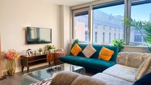 Un salon avec un canapé vert, un téléviseur à écran plat, une table basse et une grande fenêtre donnant sur des immeubles.