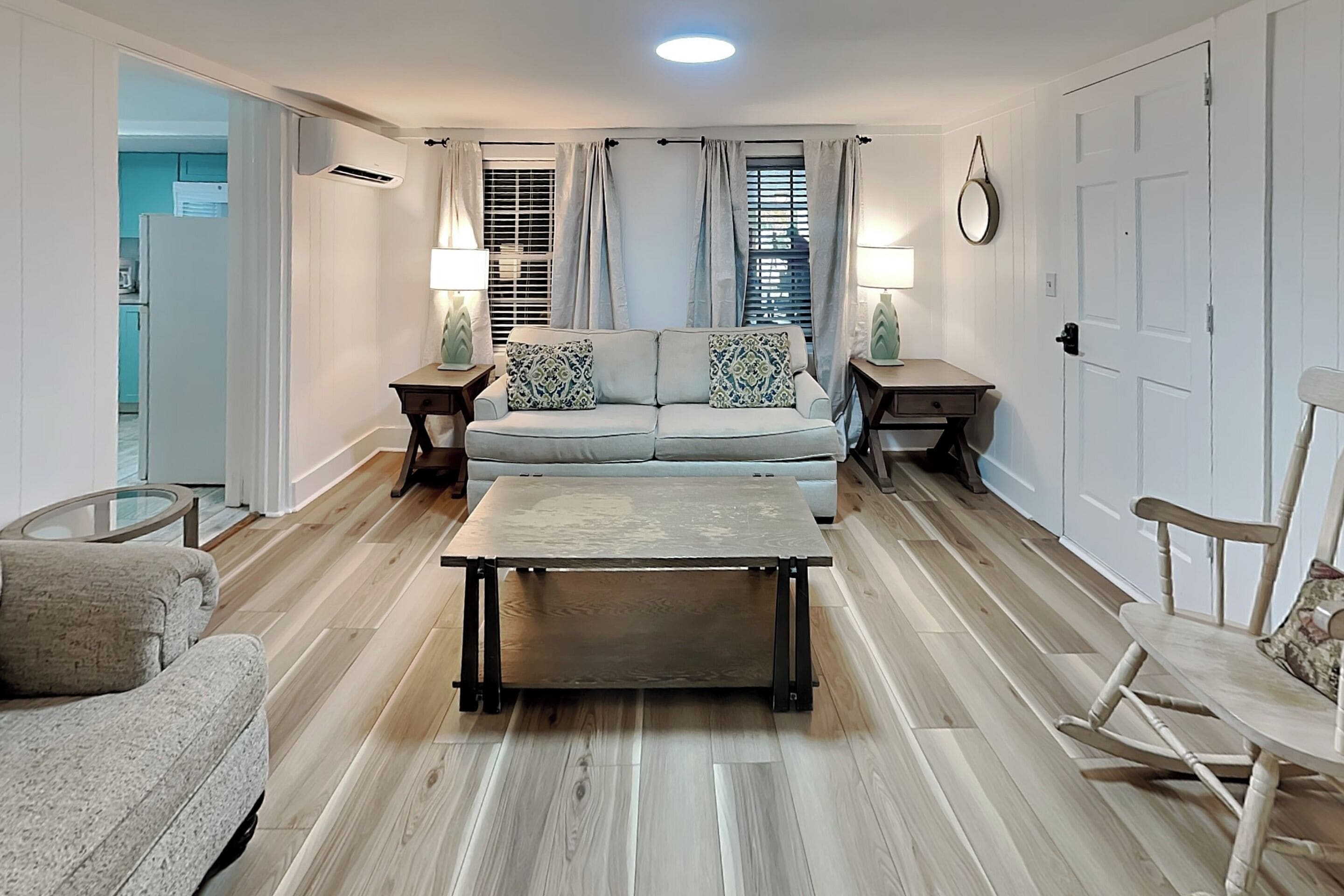 A living room with a sofa, coffee table, and chairs, a dining table, and a kitchen area in the background.