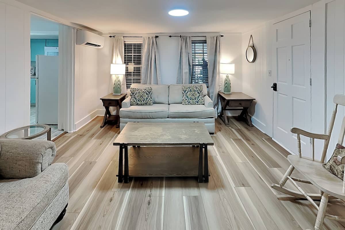 A living room with a sofa, coffee table, and chairs, a dining table, and a kitchen area in the background.