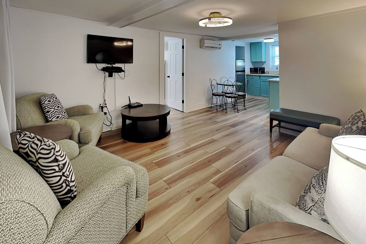A living room with a sofa, a round coffee table, a TV mounted on the wall, and a door leading to a kitchen area.