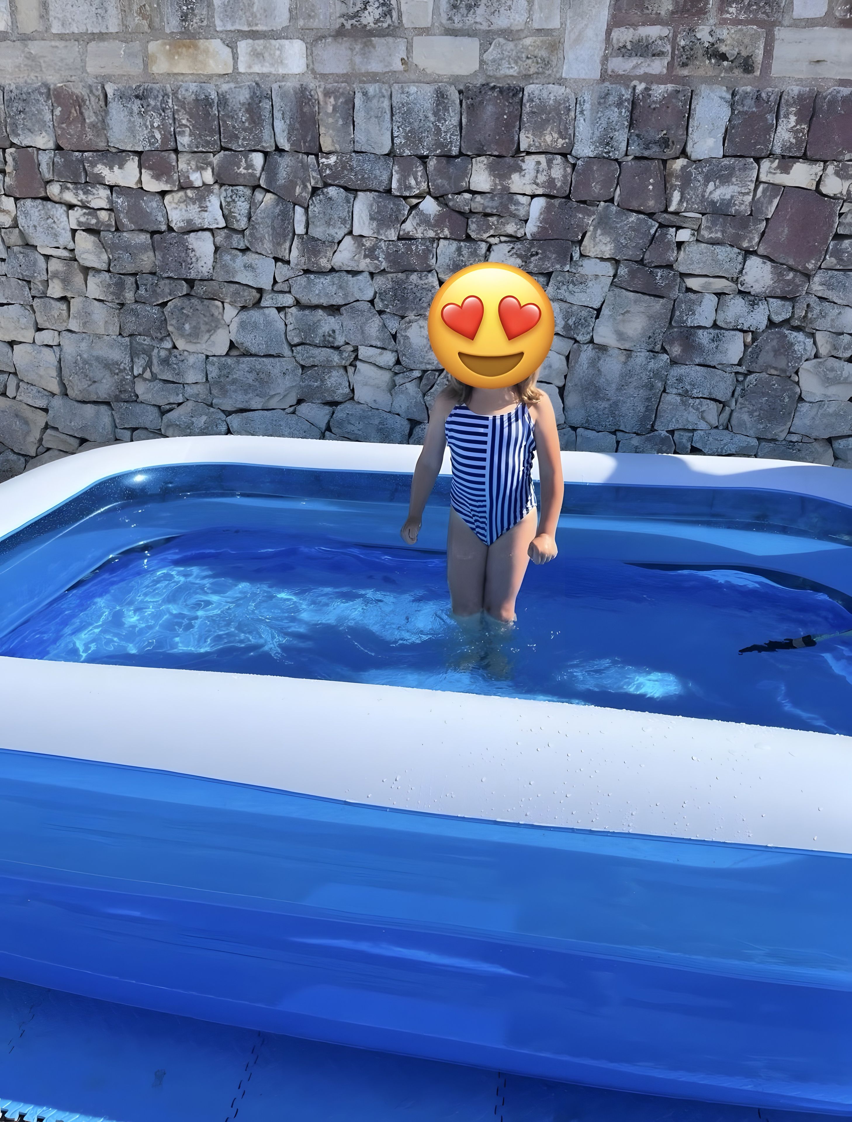 Et barn i badetøj med striber står i en lille pool med en stenvæg i baggrunden.