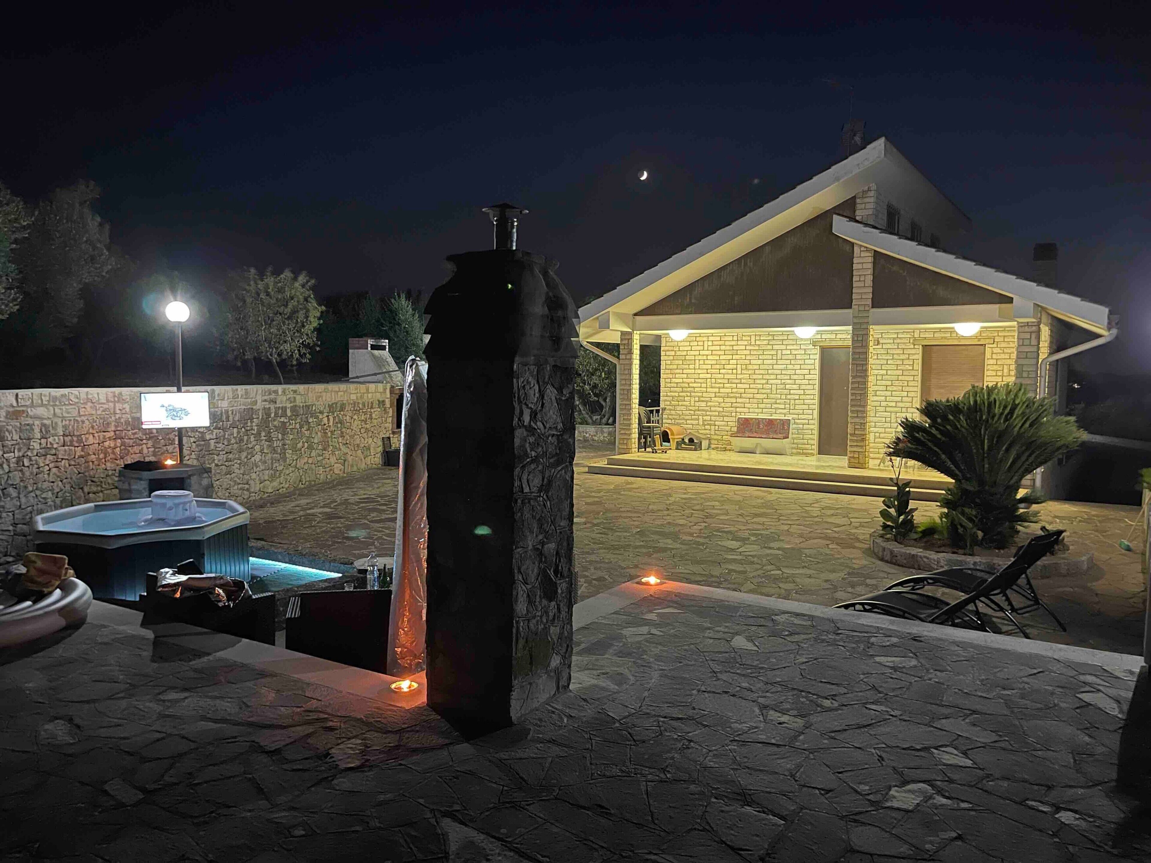 A stone patio with a hot tub, a stone pillar, and a covered outdoor seating area with a brick wall and a wooden door.