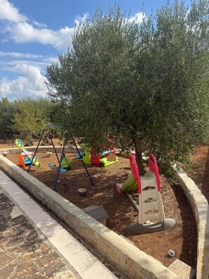 Un'area giochi con altalene e scivoli, circondata da alberi e pacciamatura.