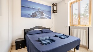 Une chambre à coucher comprenant un lit, une table de chevet, une étagère fixée au mur, une fenêtre donnant sur un paysage agréable et un tableau représentant une scène côtière.