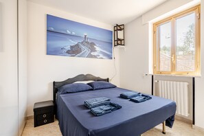 A bedroom with a bed, a nightstand, a wall-mounted shelf, a window with a view, and a painting of a coastal scene. - Happy's Casa: Experience the Countryside 20 Minutes from the Sea (Putignano)