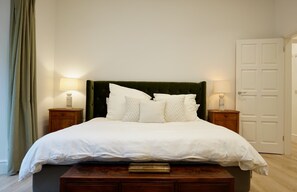 A neatly made bed with a green upholstered headboard, two bedside tables with lamps, and a wooden nightstand with a tray. - Spacious 1BDR Garden Flat in Charlton (London)