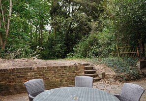 Une terrasse avec des chaises et une table en osier, entourée d’un mur de briques et d’une clôture en bois, et un luxuriant feuillage en arrière-plan.