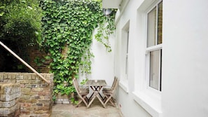 A white building with a brick wall partially covered by green ivy. There is a wooden table and two chairs outside. - Spacious 1BDR Garden Flat in Charlton (London)