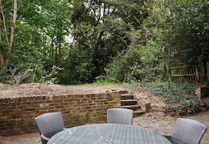 A patio with wicker chairs and a table, surrounded by a brick wall and a wooden fence, with dense greenery in the background. - Spacious 1BDR Garden Flat in Charlton (London)