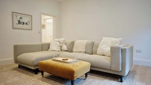 A living room with a sofa, ottoman, and a framed picture on the wall. - Spacious 1BDR Garden Flat in Charlton (London)
