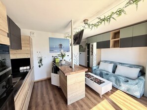 Un espace de vie compact comprenant un coin cuisine, un canapé, une table basse et un tableau représentant une jetée.