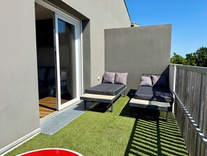 Un balcon dotĂ© dâune porte-fenĂȘtre coulissante, de deux fauteuils confortables avec coussins et dâun revĂȘtement de sol en gazon synthĂ©tique.