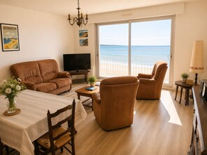A living room with a sofa, armchair, TV, and a view of the sea through a large sliding door. - Sea & Pines Apt with Terrace (La Baule-Escoublac)