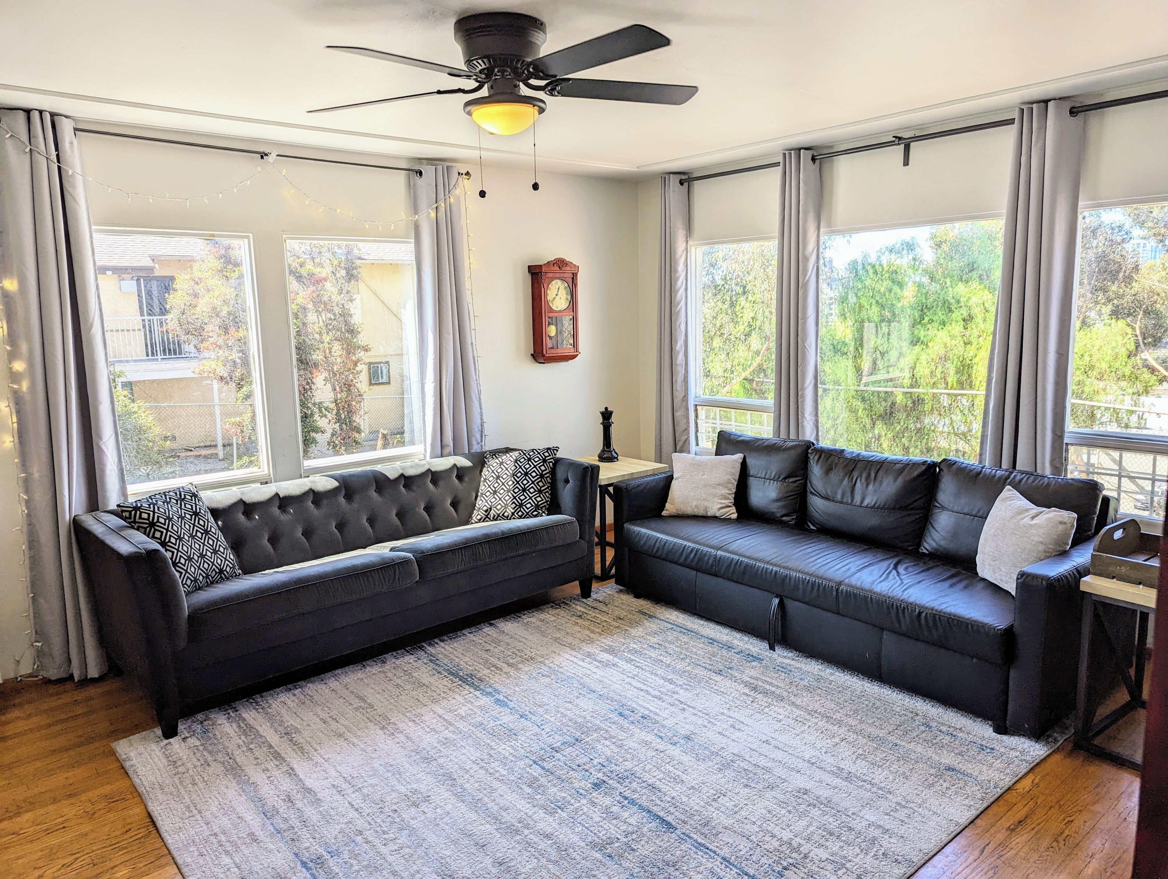 Ein Zimmer mit zwei schwarzen Ledersofas, einem Deckenventilator, einer Uhr und großen Fenstern mit Vorhängen.