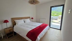 A bedroom with a bed, a red throw, a nightstand, a lamp, a radiator, and a balcony with a view. - Bright, new apartment with a terrace 3* (Briançon)