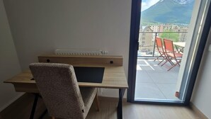 A room with a desk, chair, radiator, and a view of a balcony with chairs and a mountain. - Bright, new apartment with a terrace 3* (Briançon)