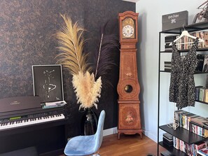 Ein Zimmer mit einem Klavier, einer hohen Holz-Uhr, einem blauen Sessel, einem Bücherregal voller Bücher und einem gerahmten Bild an der Wand.