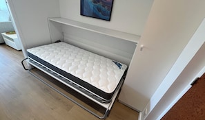 Ein kleines Bett mit Matratze und Metallgestell, an einer Wand platziert, oberhalb befindet sich ein Regal.