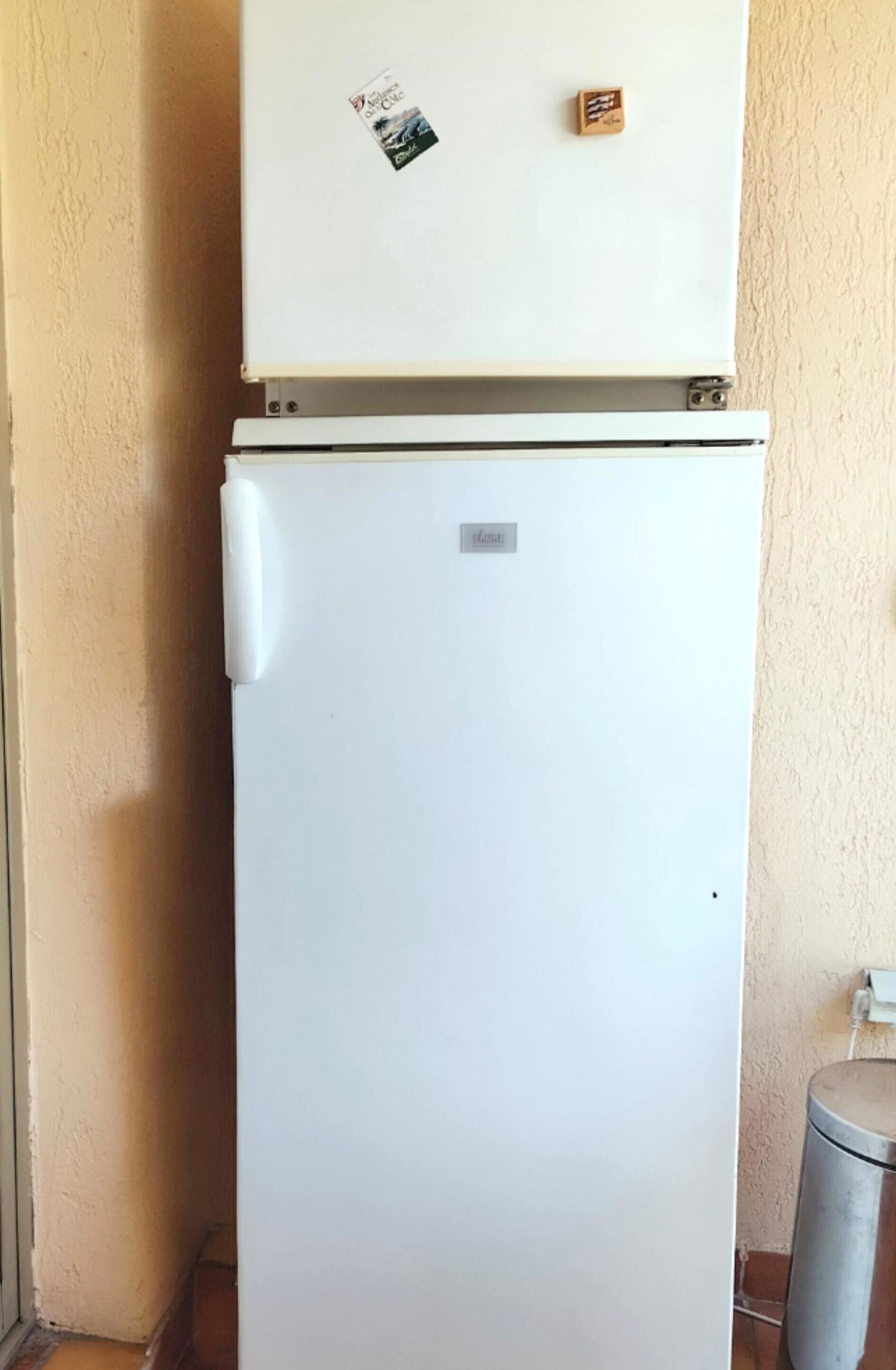 A white refrigerator with a small sticker and a metal plate on top, located in a corner with a beige wall and a trash can nearby.