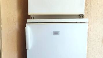 A white refrigerator with a small sticker and a metal plate on top, located in a corner with a beige wall and a trash can nearby.