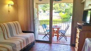 Et værelse med sofa, et bord med stole, et fjernsyn og en balkon med udsigt over grønne omgivelser.