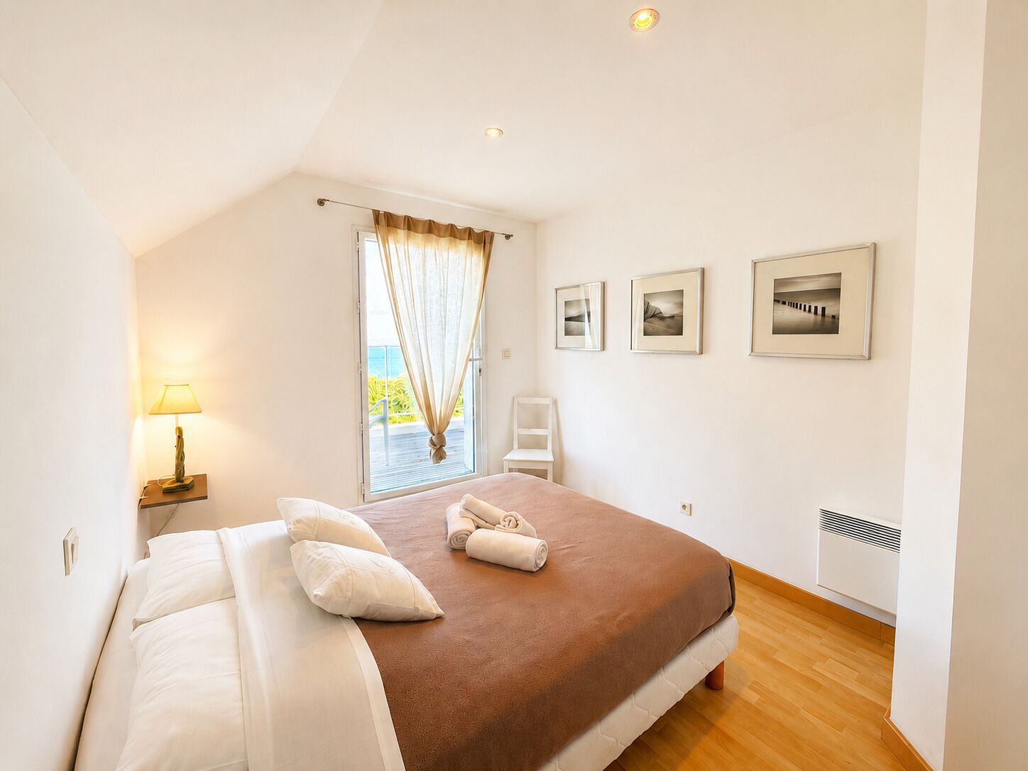 A bedroom with a bed, a nightstand with a lamp, a window with curtains, and three framed pictures on the wall.