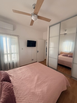 Ein Schlafzimmer mit Bett, Deckenventilator, Fernseher, einer Schiebetür und einem Fenster mit Jalousien.