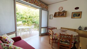 A room with a sofa, a dining table with chairs, a whiteboard, and a view of the outdoors. - Cozy Apartment With Terrace in Central Lignano (Lignano Sabbiadoro)