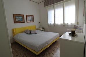 A bedroom with a bed, a desk, a chair, a nightstand, and two framed pictures on the wall. - Cozy Apartment With Terrace in Central Lignano (Lignano Sabbiadoro)