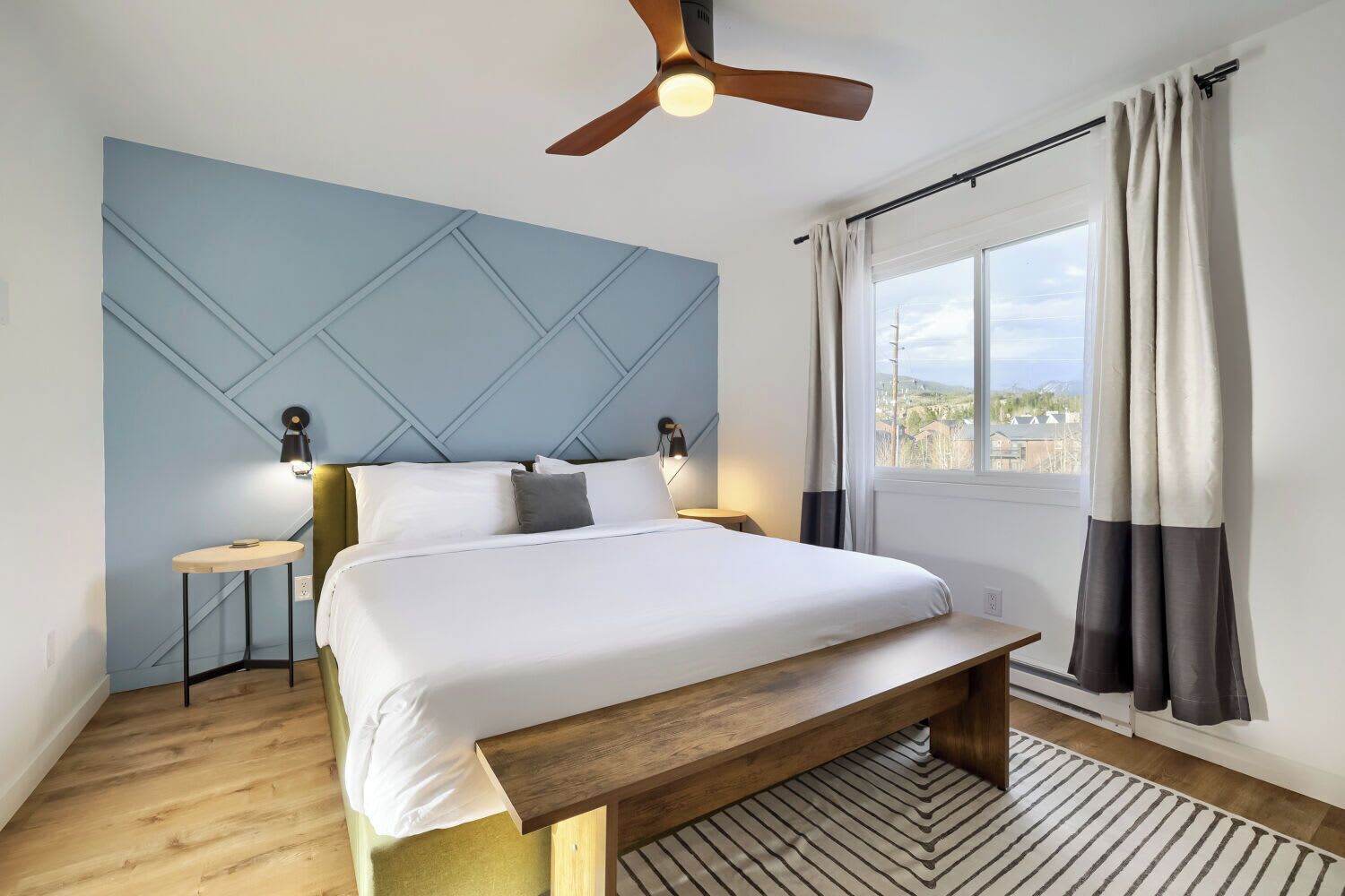 A bedroom with a bed, bedside tables, a ceiling fan, a window with curtains, and a view of the outside.