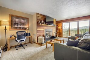 Ein gemütliches Wohnzimmer mit Kamin, einem großen Fenster mit Bergblick, einer bequemen Couch und einem Schreibtisch mit Stuhl.
