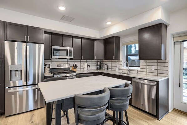 A modern kitchen with stainless steel appliances, dark cabinetry, a large island with bar stools, and a window providing natural light. - Modern Mountain Living, New Listing (Silverthorne)