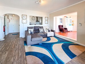 Ein Wohnzimmer mit einem Ecksofa, einem gemusterten Teppich und einer durch einen Rundbogen sichtbaren Küche.