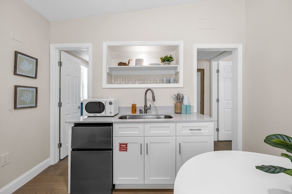 Cocina moderna con gabinetes blancos, un refrigerador de acero inoxidable, un microondas y un lavamanos con grifo. Hay cuadros en la pared y una repisa sobre el lavamanos con vasos y una planta.