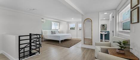 A spacious room with a bed, a small seating area, a mirror, and a kitchenette.